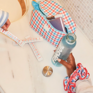 A lifestyle shot of a person holding a light blue glass bottle with 'FINNSUL' printed on it, next to a red, white, and blue gingham zippered pouch and a matching scrunchie on a white marble countertop.